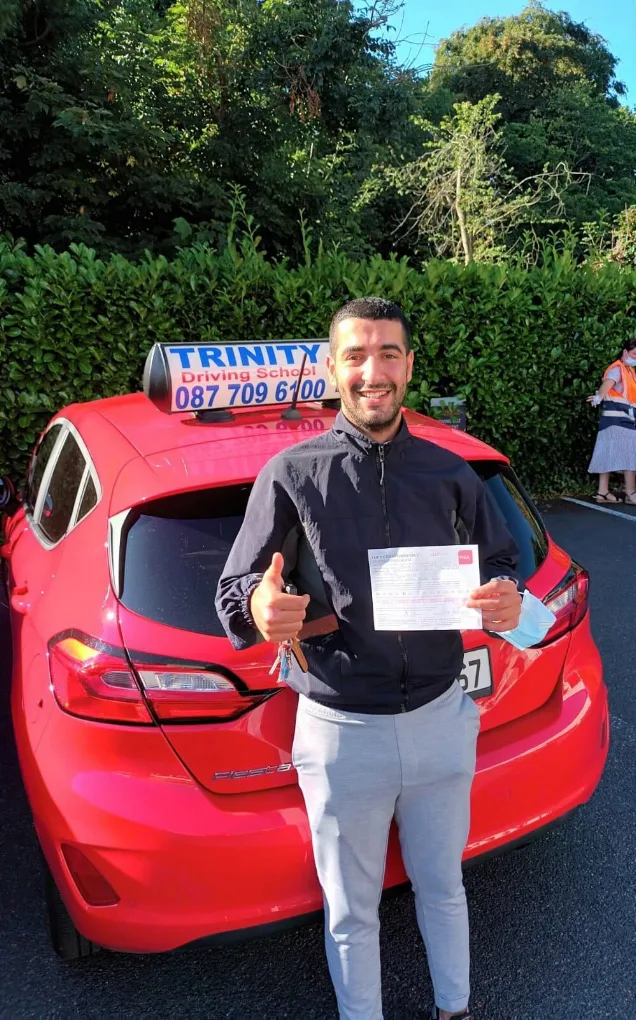 A Trinity Driving School student holding their driving test pass certificate beside the school car, with the Trinity Driving School roof sign showing 087 709 6100
